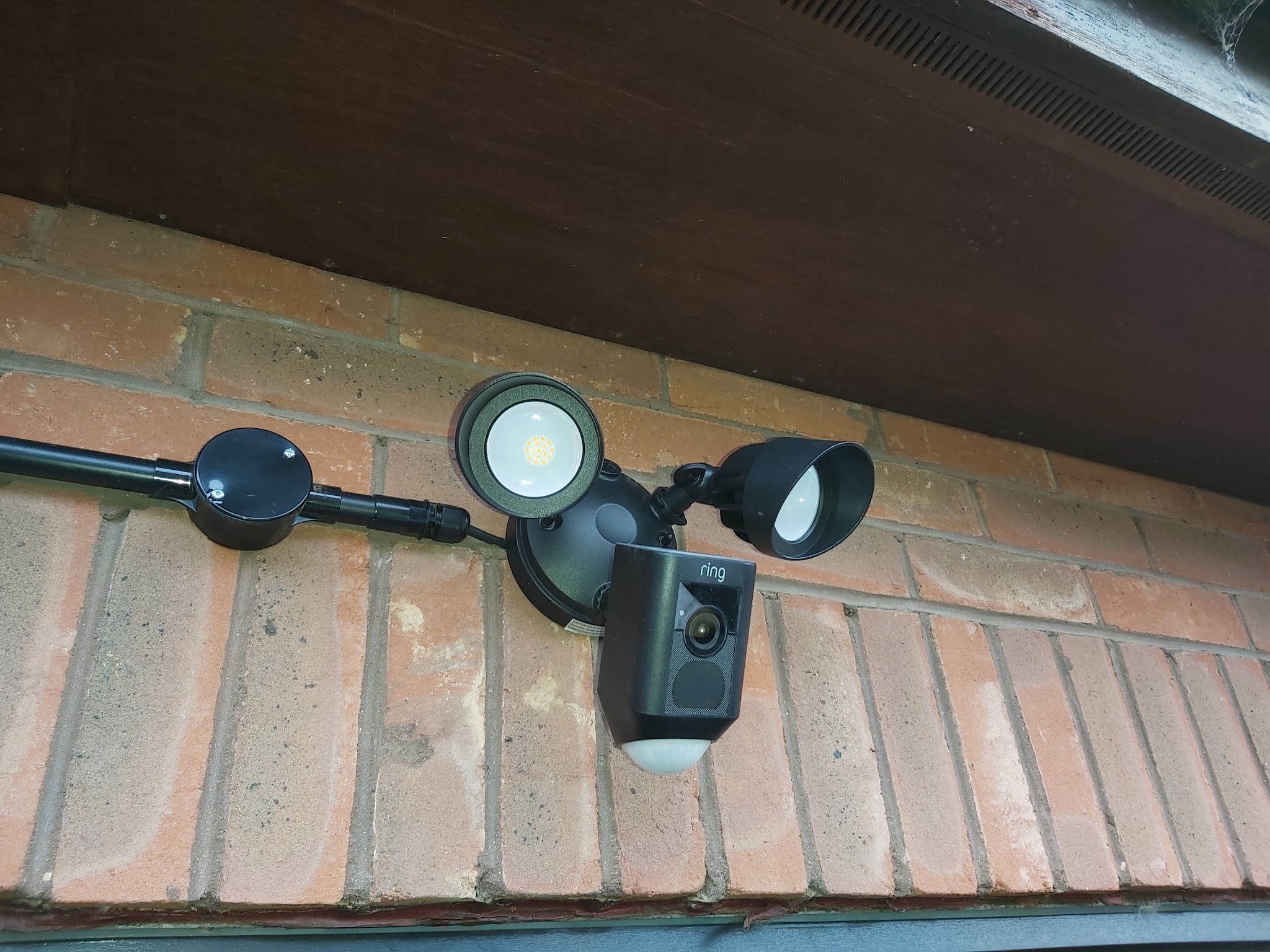 A floodlight security camera fitted under a soffit on a brick wall
